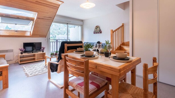 Een gezellige, goed verlichte kamer met een houten eettafel gedekt voor vier personen, omringd door houten stoelen. Een televisie staat op een lage houten standaard, en er is een zithoek met een bank en een fauteuil. De kamer heeft een groot raam met uitzicht op het buitenleven, en een houten plafond met een dakraam.