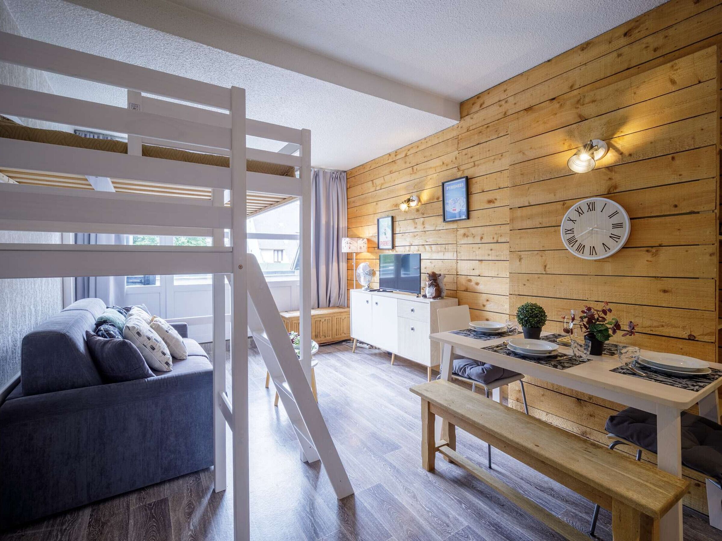 Ett modernt vardagsrum med en sovplats på entresolplan, en matplats med ett träbord och en bänk, en soffa och en tv.