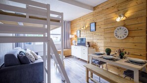 Un espai de vida modern amb un llit d'entreplà, una zona de dinar amb taula i panxa de fusta, un sofà i una televisió.