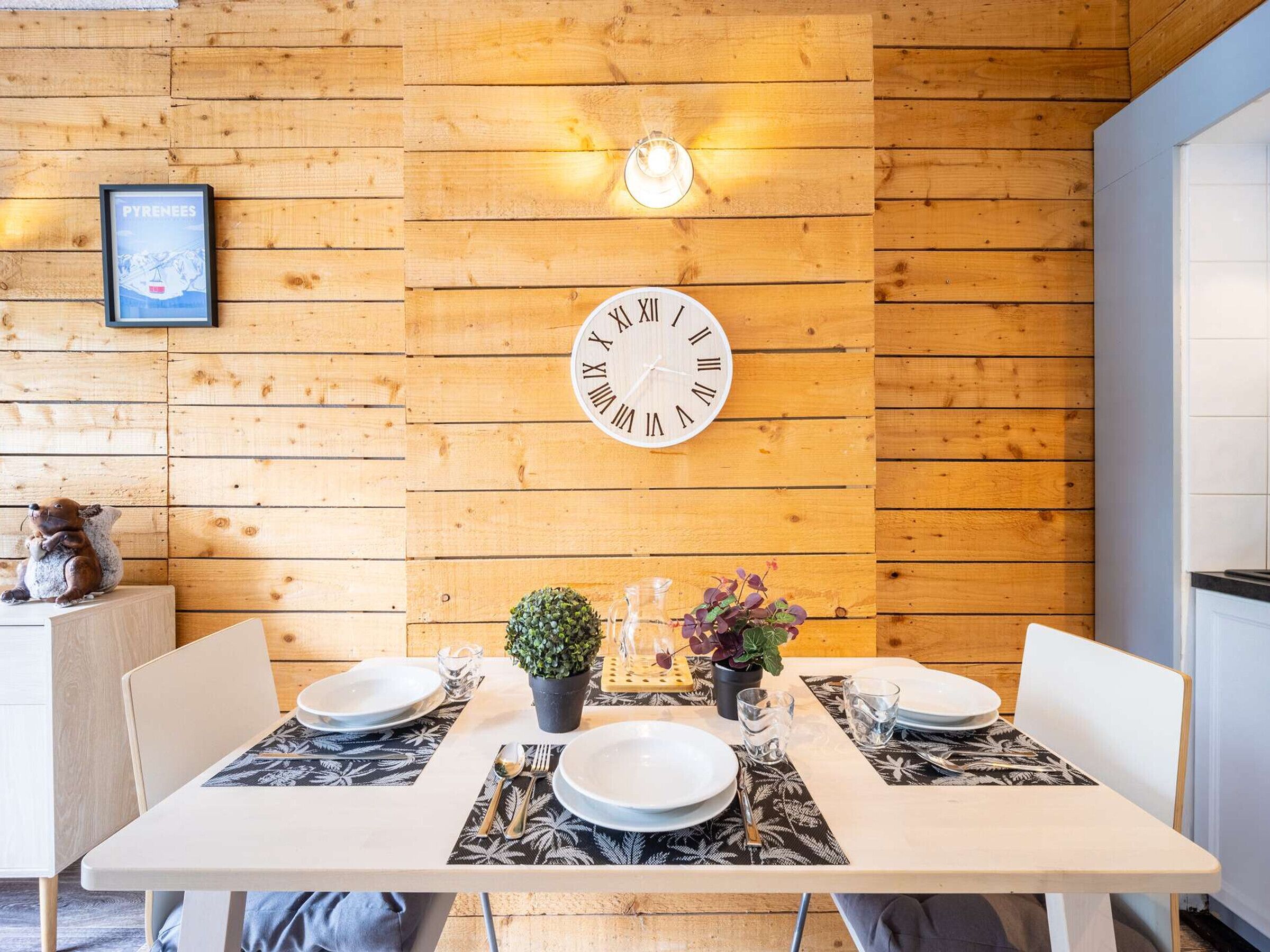 A wooden interior with a wall-mounted clock, a framed picture, and a table set for two with plates, cutlery, and small plants.