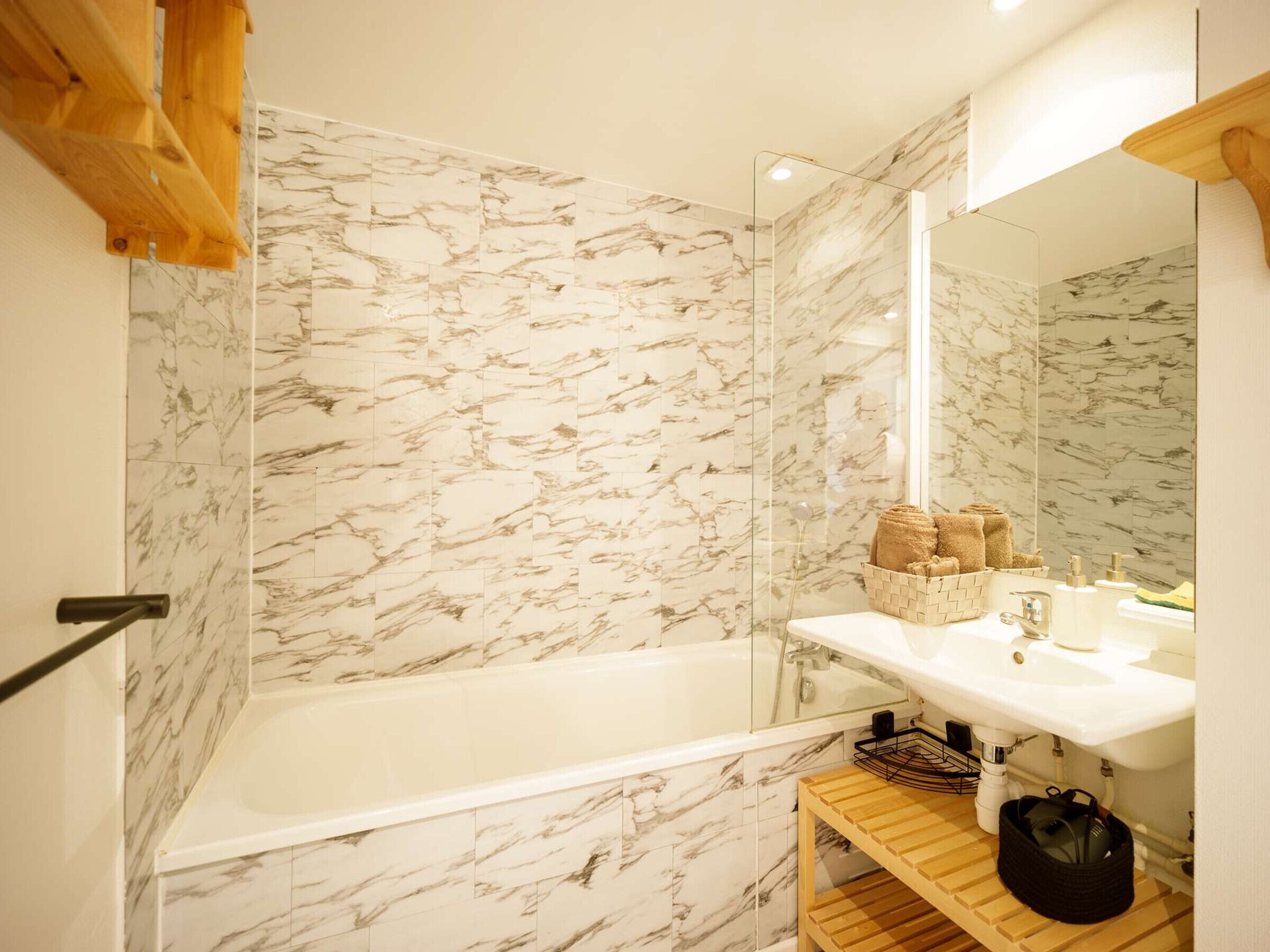 A bathroom with a marble wall, a bathtub, a wooden shelf, and a mirror.