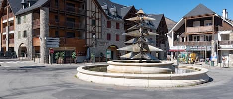 Ett offentligt torg med en central fontän, omgivet av traditionella trähus med balkonger och skyltar som visar vilka lokala företag som finns i närheten.
