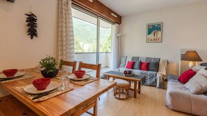 A cozy living room with a wooden dining table, a sofa with patterned cushions, and a large window offering a view of greenery. - 4-Person Apartment in Saint-Lary with Parking (Saint-Lary-Soulan)