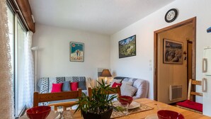 A cozy living room with a wooden dining table set for four, a sofa with patterned cushions, a potted plant, and a poster on the wall. - 4-Person Apartment in Saint-Lary with Parking (Saint-Lary-Soulan)