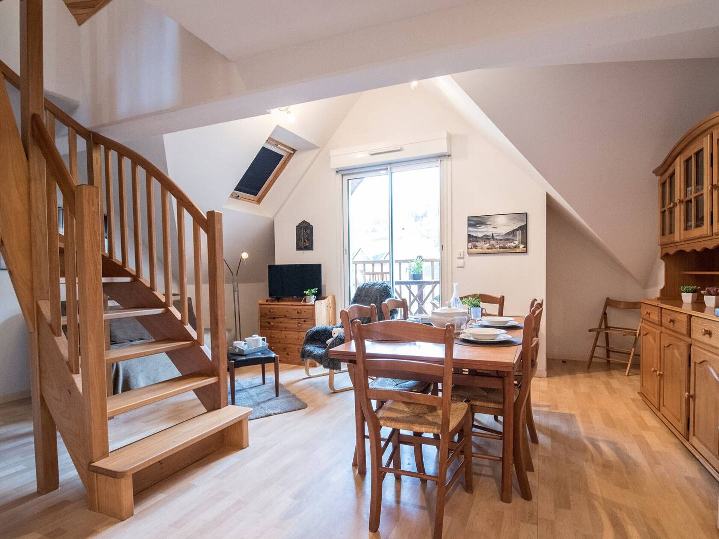 Ein großzügiges Loft mit einer Holztreppe, einem Essbereich mit Holztischen und -stühlen sowie einem großen Fenster, das für natürliches Licht sorgt.
