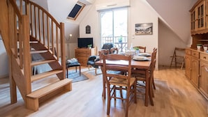 A spacious loft with a wooden staircase, a dining area with wooden chairs and a table, and a large window providing natural light.