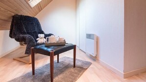 A cozy room with a sloped ceiling, a window, a radiator, a plush chair, a small table with a cup and saucer, and a teddy bear.