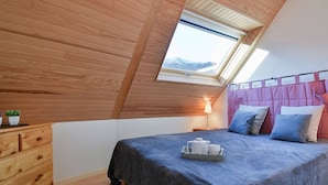 A bedroom with a sloped ceiling, a skylight, a bed with pillows, a bedside table with a lamp, and a dresser.