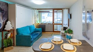 Un salon moderne avec un canapé de couleur turquoise, une table à manger pour quatre personnes et une grande fenêtre donnant sur des immeubles.