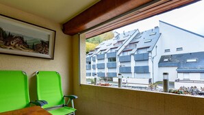 Un balcon avec une table en bois et deux chaises vertes, donnant sur un immeuble moderne doté de nombreux balcons.
