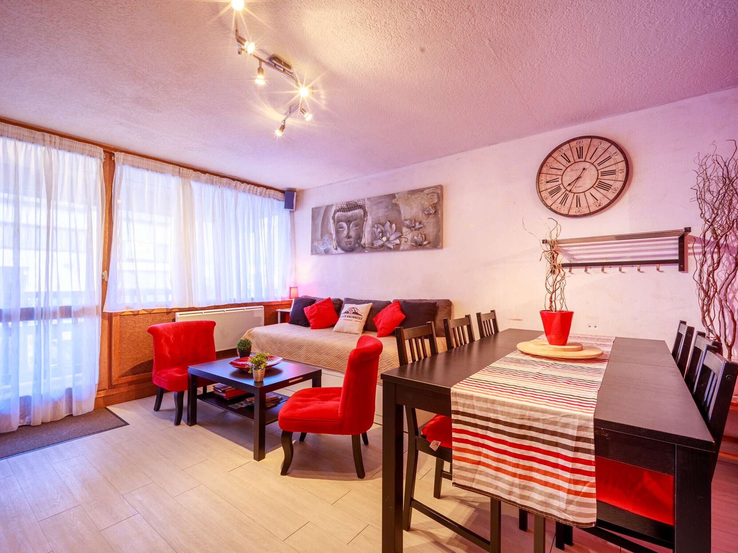 Un salon moderne avec un canapé, des fauteuils rouges, une table à manger recouverte d ’une nappe à rayures, une horloge au mur et un tableau représentant un Bouddha.
