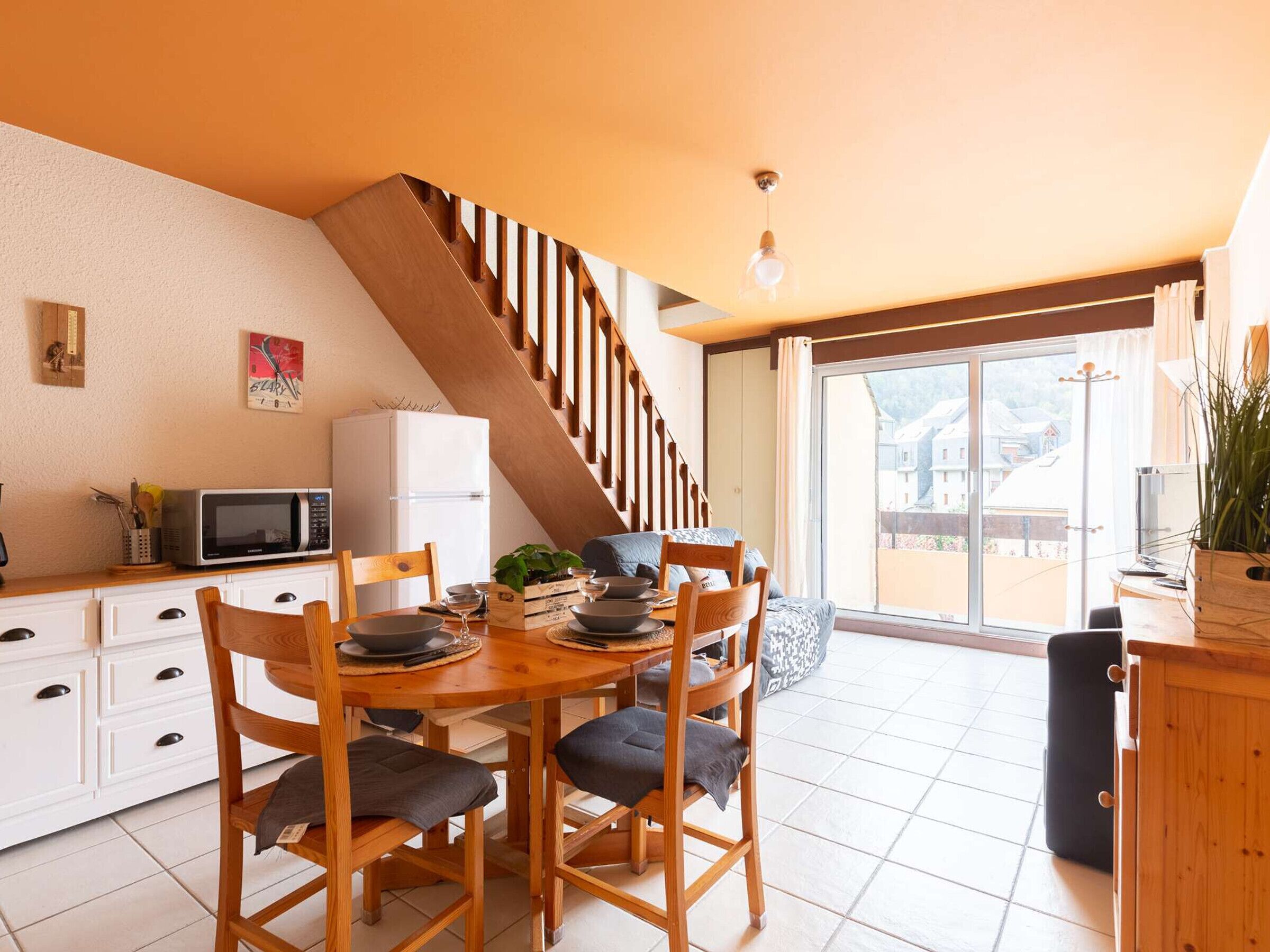 Un comedor con mesa y sillas de madera, una cocina con microondas y refrigerador, y una escalera que lleva a un balcón con vistas a casas.