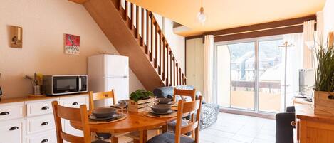 Un comedor con mesa y sillas de madera, una cocina con microondas y refrigerador, y una escalera que lleva a un balcón con vistas a casas.