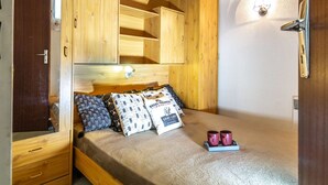 Une chambre avec une armoire en bois, un lit agrémenté d’oreillers à motifs et une petite table sur laquelle sont posées deux tasses.