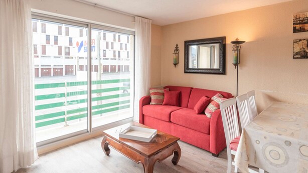 A living room with a red sofa, a wooden coffee table, and a view of a building through large windows. - Apartment for 6 people close to ski slopes (Saint-Lary-Soulan)