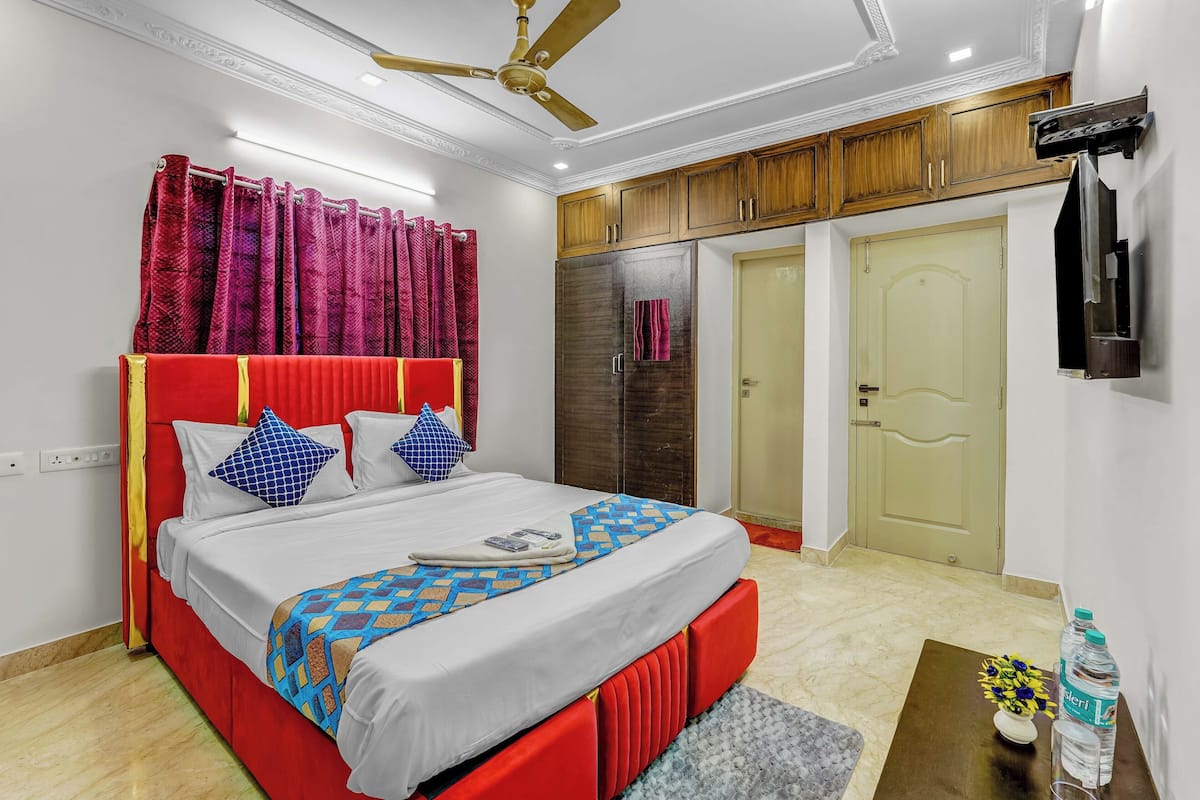 A bedroom with a red bed, a ceiling fan, wooden cabinets, a wardrobe, and a small table with a vase and water bottles.