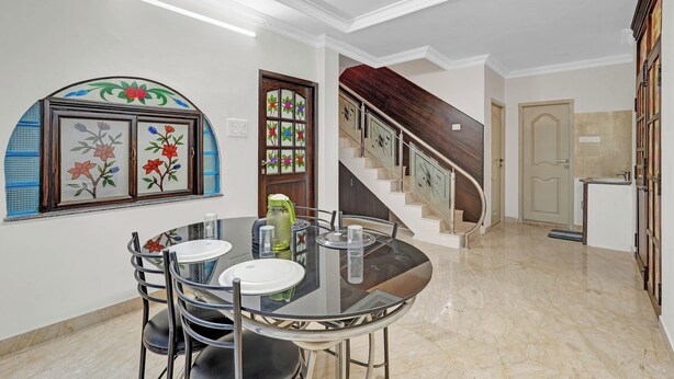 A dining area with a round table, chairs, and a glass top. There is a staircase with a wooden railing leading to another floor. The walls are white, and there is a door with a stained glass window. - Butterfly Residency (Chennai)