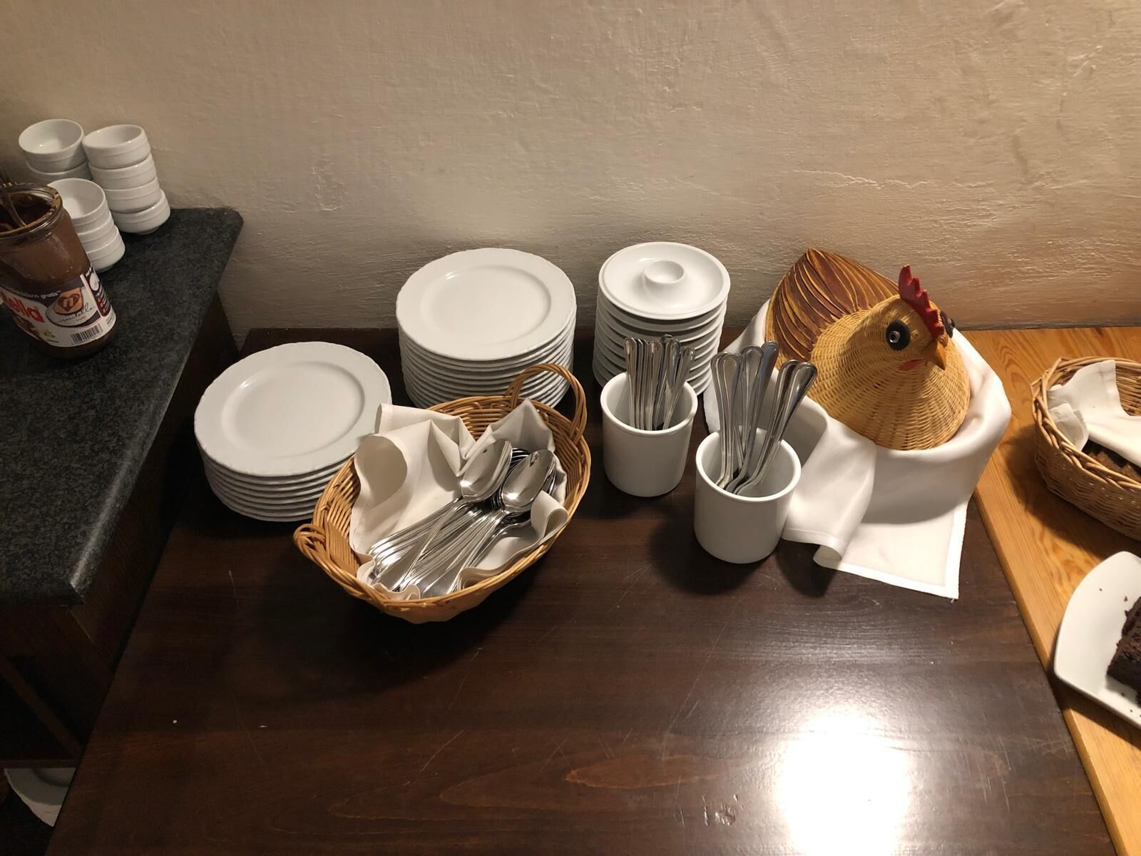 Una mesa dispuesta con platos blancos, cuencos apilados y cubiertos en cestas y tazas.
