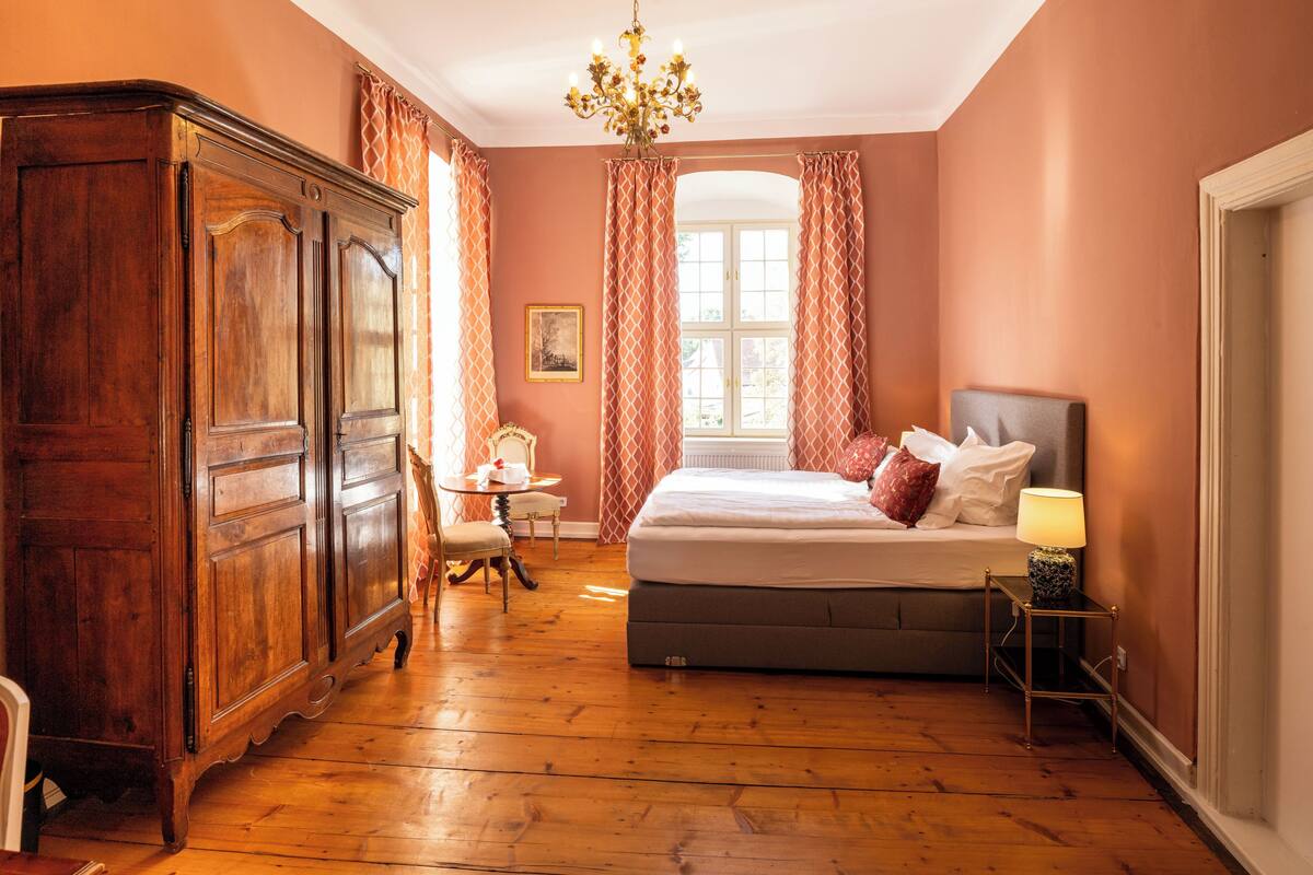 A bedroom with a large wooden wardrobe, a bed with pillows, a small table, and a lamp.