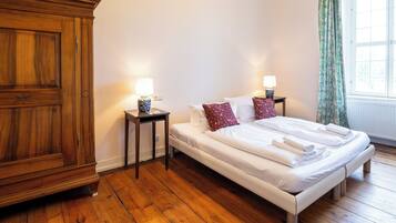Quarto com guarda-roupa de madeira, cama com roupa de cama branca, duas mesas de cabeceira com abajures e janela com cortinas.