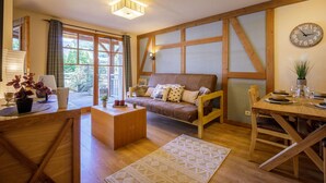 Un salón acogedor con un sofá de madera, una mesa de comedor para dos personas y un amplio ventanal con cortinas.