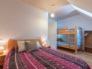 Une chambre avec un lit superposé, un grand lit recouvert d’une couvre-lit à motifs, une table de chevet avec une lampe et une armoire en bois.