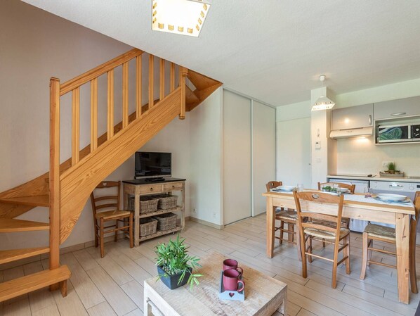 Un espace de vie moderne avec un escalier en bois, un coin repas avec des meubles en bois, une cuisine avec des placards blancs et un téléviseur à écran plat posé sur un meuble en bois.