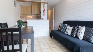 Een compacte woonruimte met een eethoek, een bank met gestreepte kussens en een keuken met houten kastjes en een 'BAR' bord.
