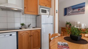 A compact kitchen with wooden cabinets, a white refrigerator, a microwave, and a sink. There's a small table with a magazine and a potted plant.