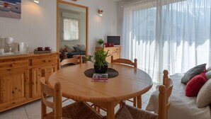 A cozy living room with a wooden dining table, chairs, a sofa, and a TV. There is a window with sheer curtains and a clock on the wall.