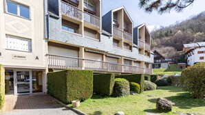 A multi-story building with a ground-level entrance, balconies, and a well-maintained garden with hedges and rocks.