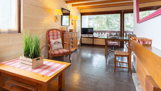 Una habitación acogedora con vigas de madera, una mesa de madera con un mantel individual, un sillón escocés, un televisor sobre un mueble y vistas al verdor exterior.