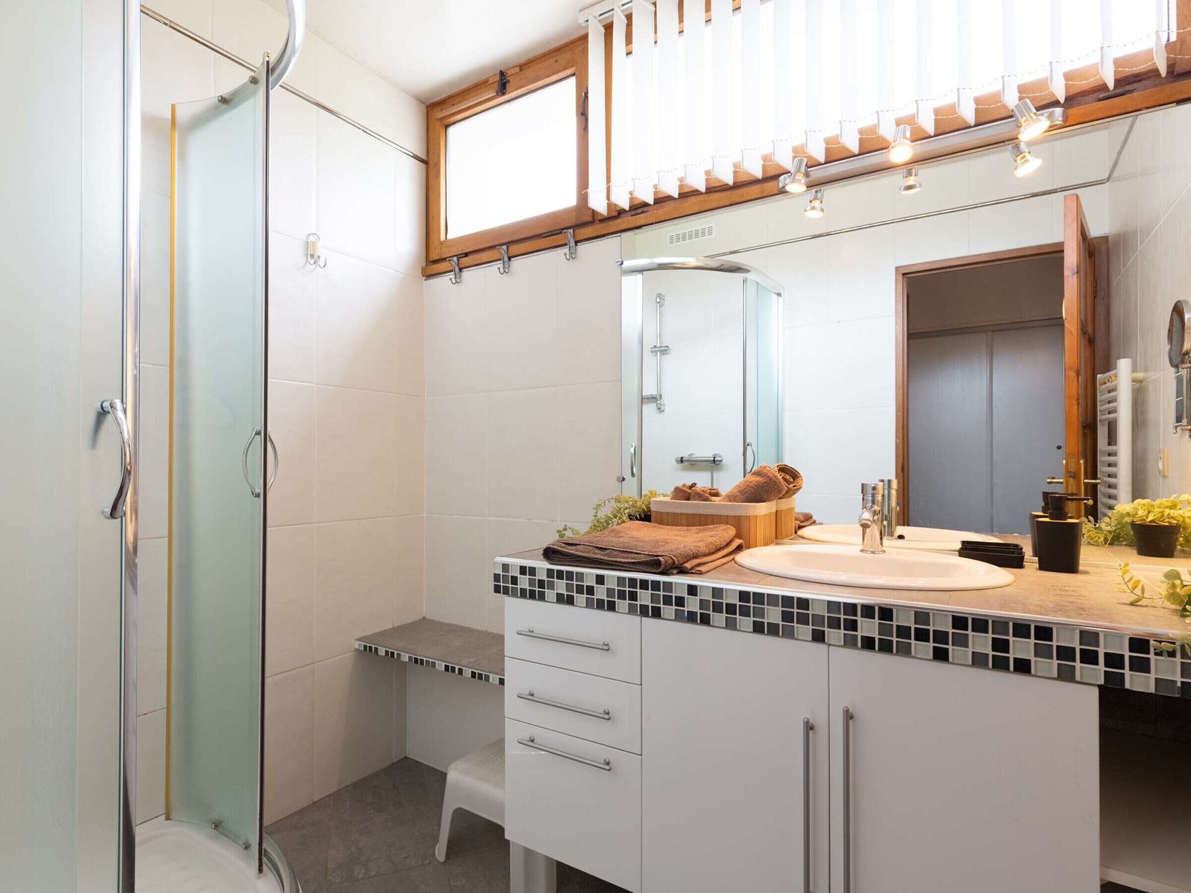 Een moderne badkamer met een douchecabine van glas, een witte wastafel met betegelde spatwand en een grote spiegel boven de wastafel.