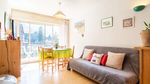 Un salon chaleureux avec un canapé gris, une table à manger avec des chaises, une grande fenêtre et un balcon.