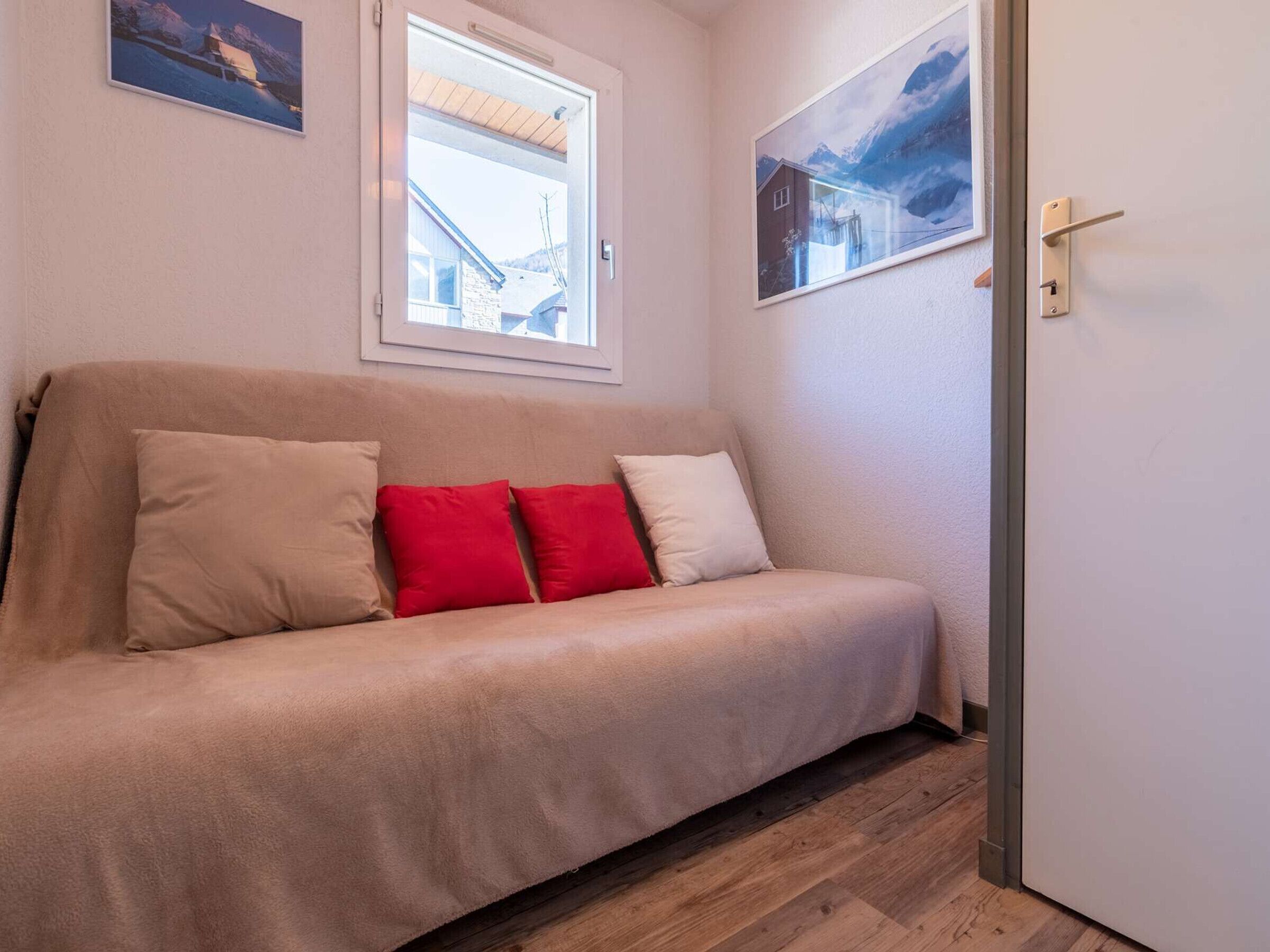 Un sofá beige con cuatro cojines en una habitación con suelo de madera, una ventana con vista a un edificio y una puerta con picaporte.