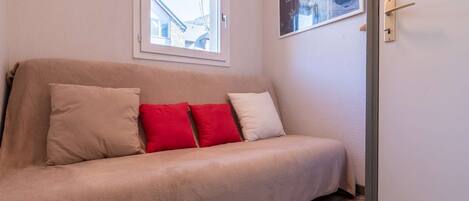 Un sofá beige con cuatro cojines en una habitación con suelo de madera, una ventana con vista a un edificio y una puerta con picaporte.