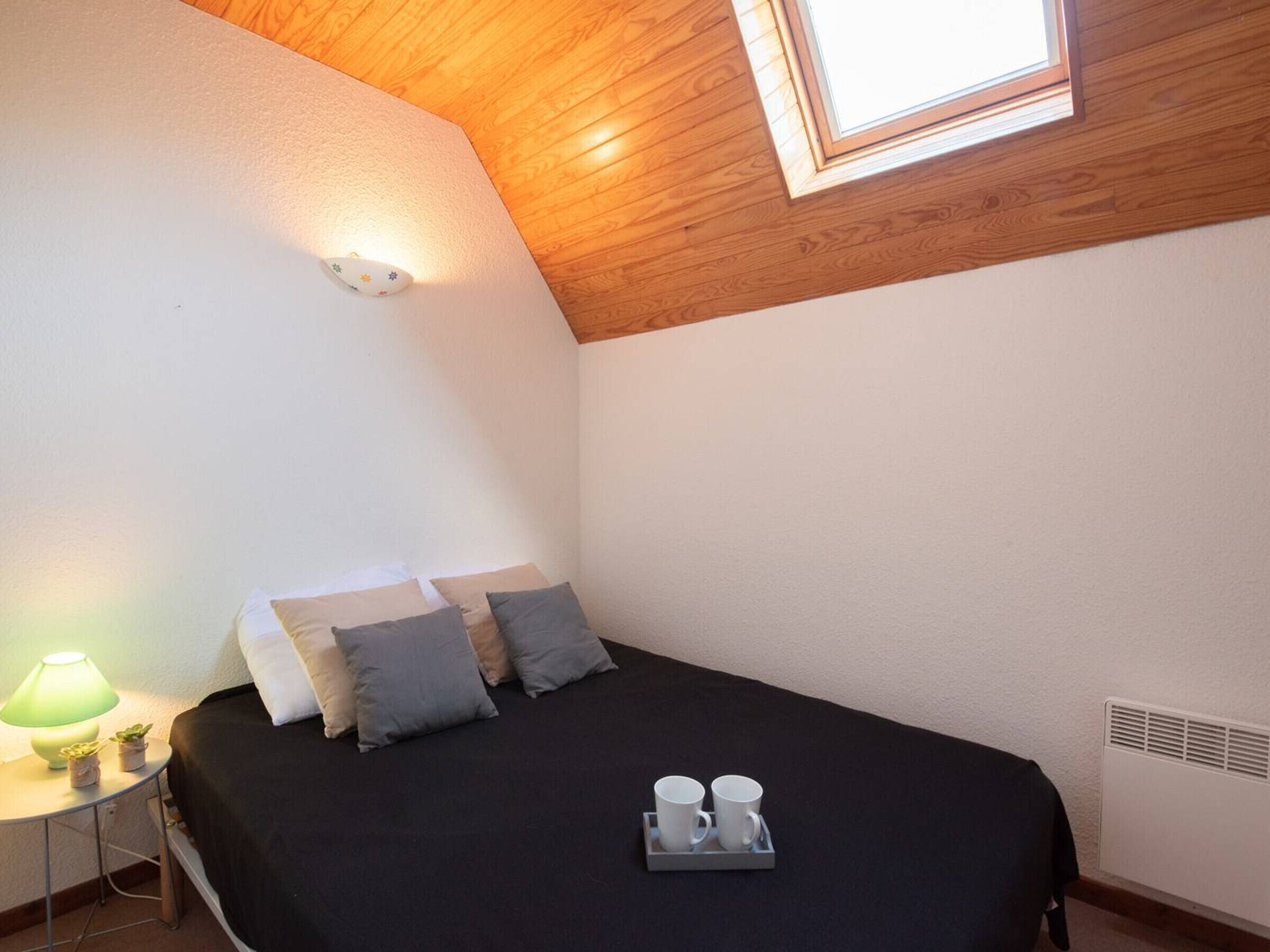A bedroom with a wooden ceiling, a skylight, a bed with pillows, a bedside table with a lamp, and a tray with cups.