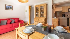 Una sala de estar acogedora con un sofá rojo, una mesa de comedor de madera dispuesta con platos y tazones blancos, un aparador con puertas de cristal, y una zona de cocina con armarios de madera y una cafetera.