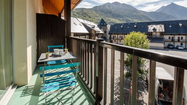 Un balcón con mesa y sillas, con vista a una calle con edificios y montañas al fondo.