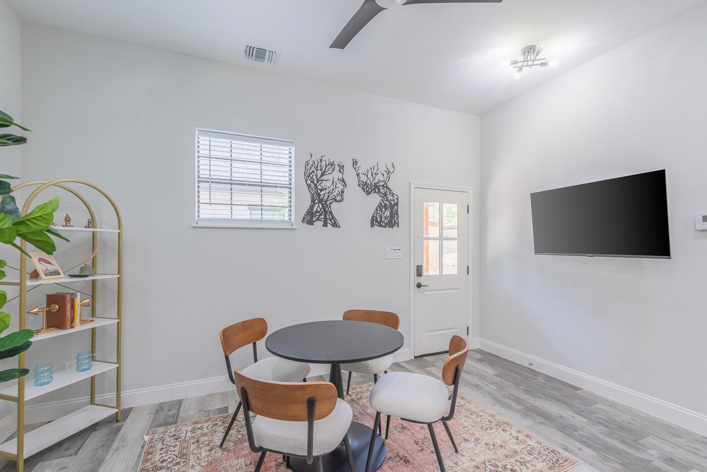 A modern living room with a round table, four chairs, a TV mounted on the wall, a bookshelf, and two windows with blinds.