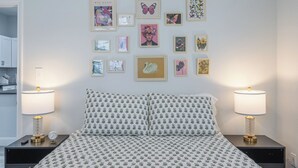Un dormitorio ordenado con una colcha estampada, dos lámparas de noche y una pared decorada con cuadros.