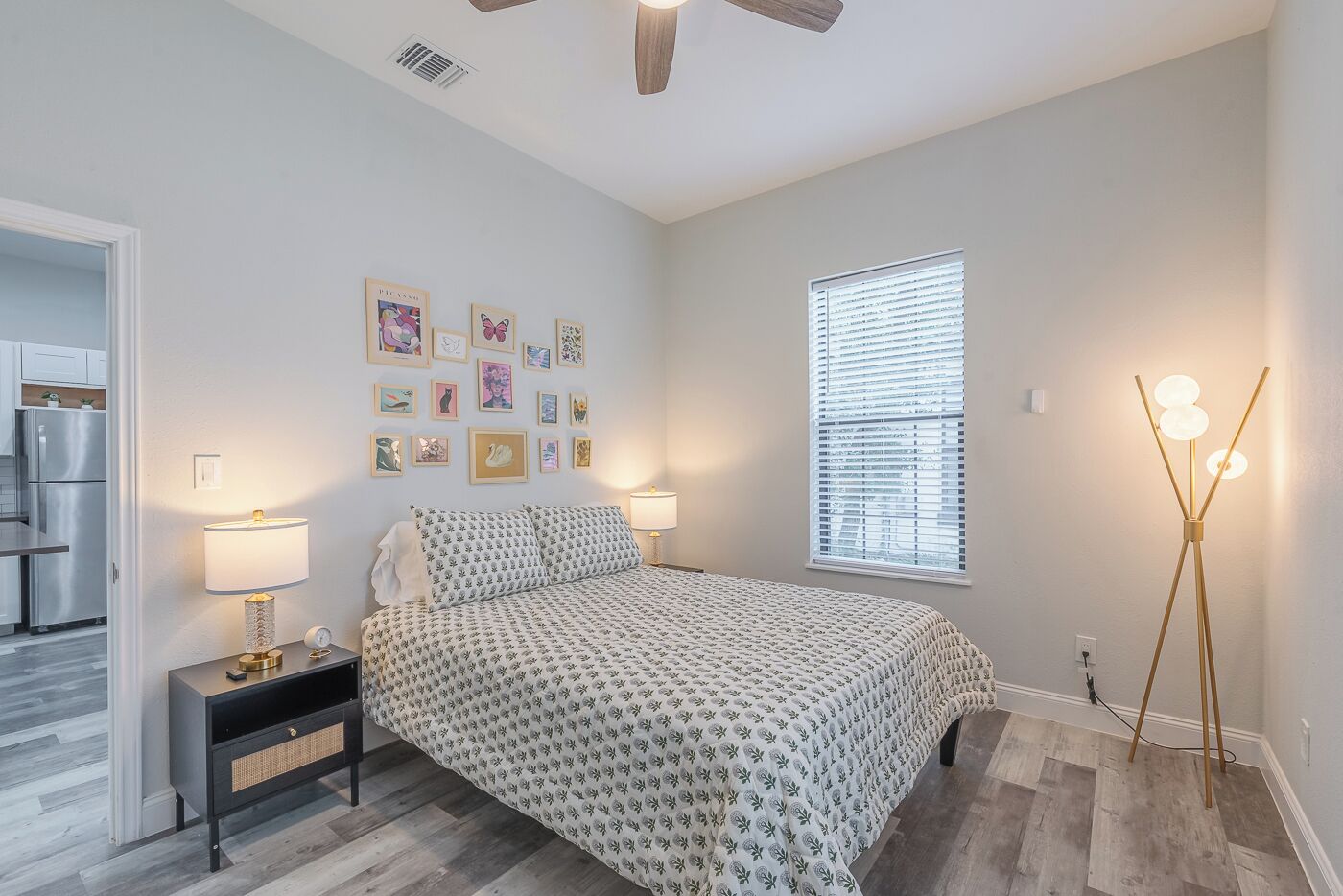 A bedroom with a bed, bedside lamps, a wall with framed pictures, a window with blinds, and a floor lamp.