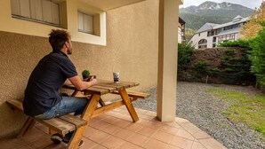 Ein Mann sitzt an einem Holztisch im Freien, auf dem ein Blumentopf und eine Tasse stehen. Im Hintergrund sind ein Berg und ein Gebäude zu sehen, und eine überdachte Terrasse bietet Schatten.