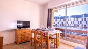A small room with a TV on a wooden dresser, a dining table with chairs, and a large window with a view of buildings and a mountain. - Cozy studio 6 pers. ski-in/ski-out (Saint-Lary-Soulan)