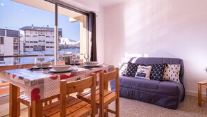 A cozy room with a dining table set for four, a purple sofa, and a large window offering a view of buildings and snow. - Cozy studio 6 pers. ski-in/ski-out (Saint-Lary-Soulan)