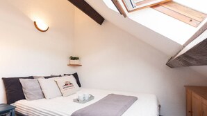 A bedroom with a sloped ceiling, a bed with pillows, a bedside table, a small shelf with a plant, and a window with a wooden frame. - Apartment for 4 in Saint-Lary-Soulan, near center and cable car (Saint-Lary-Soulan)
