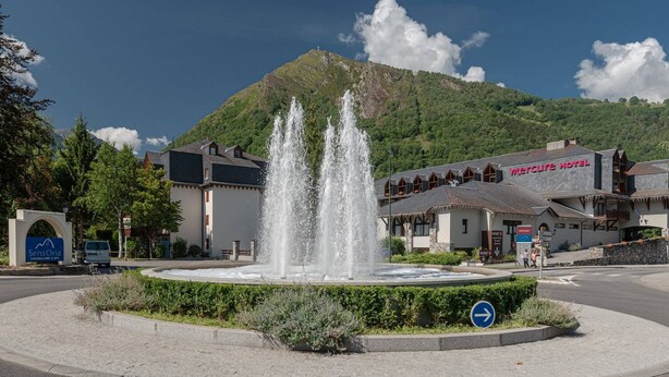 Un hotel con una fuente en el centro, rodeado de vegetación y una montaña al fondo.