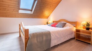 Un dormitorio con techo inclinado, tragaluz, cama con almohadas, mesita de noche con lámpara y una planta pequeña.