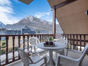Un balcon doté d’une balustrade en bois, de chaises en plastique blanc et d’une table ronde sur laquelle reposent deux tasses rouges et un pot de fleurs.