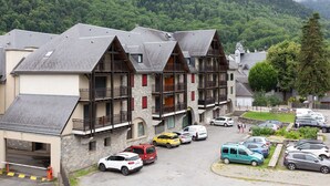 A traditional chalet-style building with a stone facade, multiple balconies, and a parking area in front.